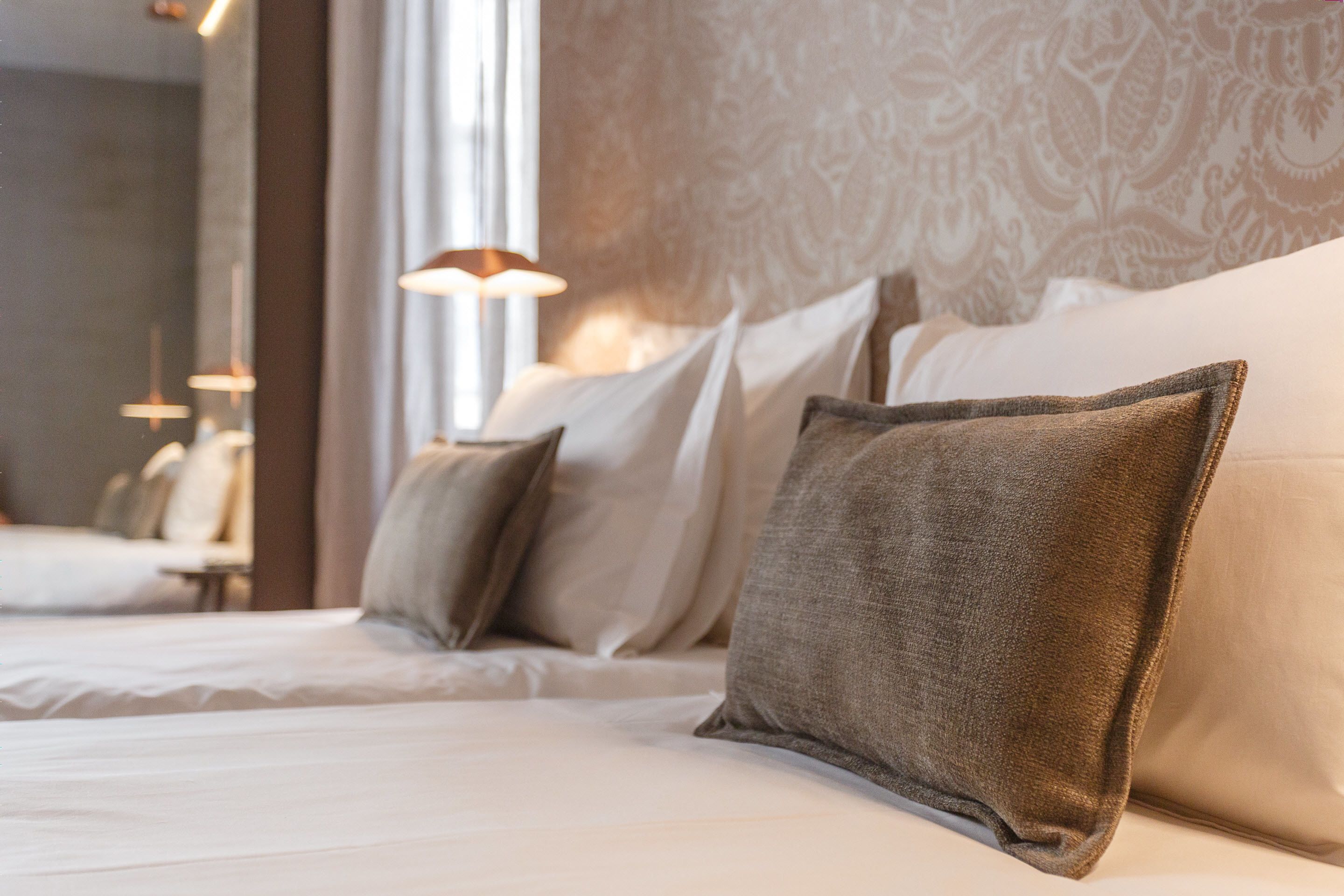 Una cama bien hecha con sábanas blancas y varios almohadones, incluyendo uno grande color marrón.