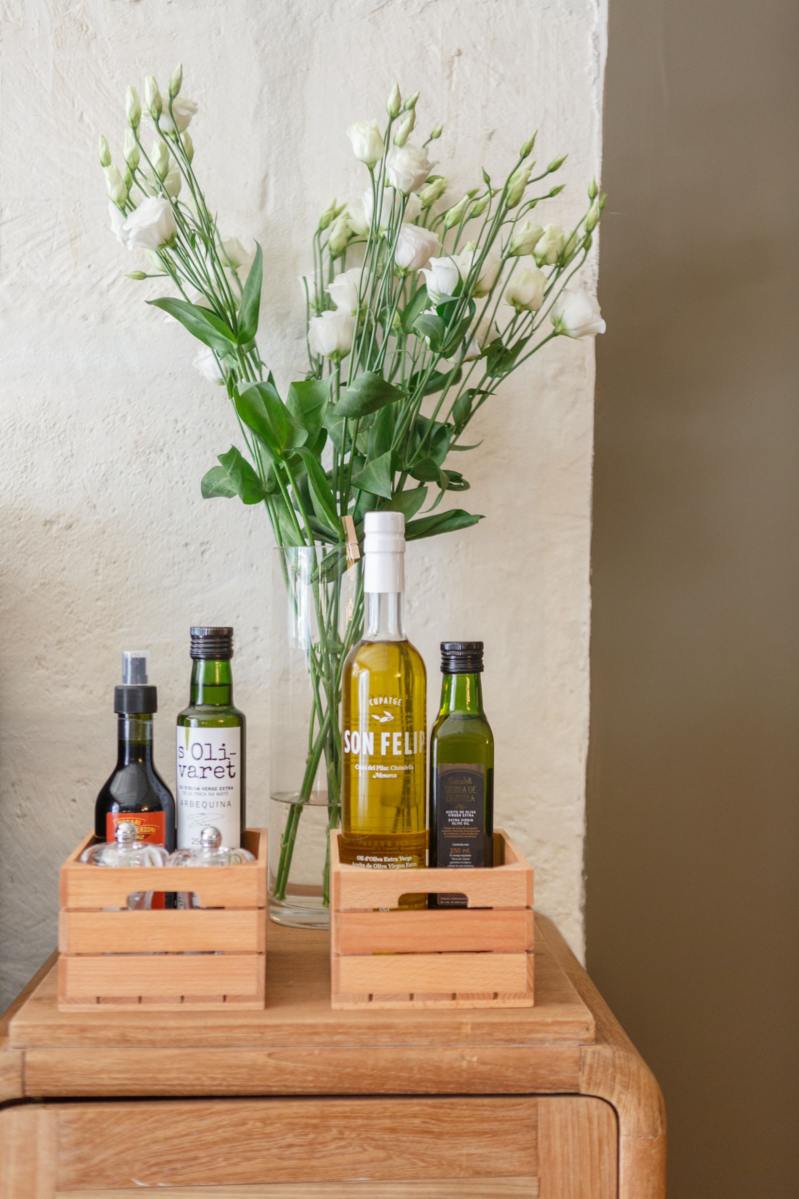 Une table en bois sur laquelle reposent deux caisses contenant des bouteilles d’huile d’olive et un vase avec des fleurs blanches.