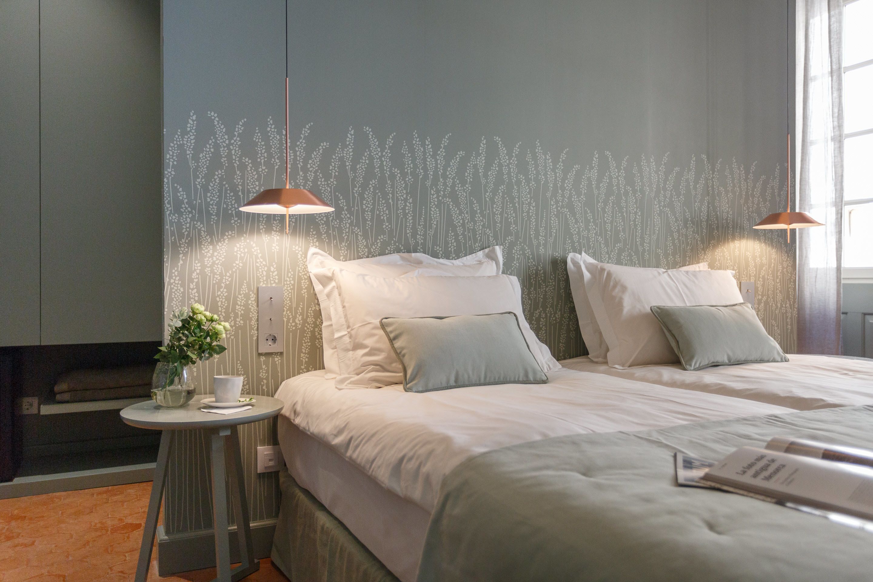 A modern bedroom with two beds, a small round table with a plant, and a wall with a floral pattern.