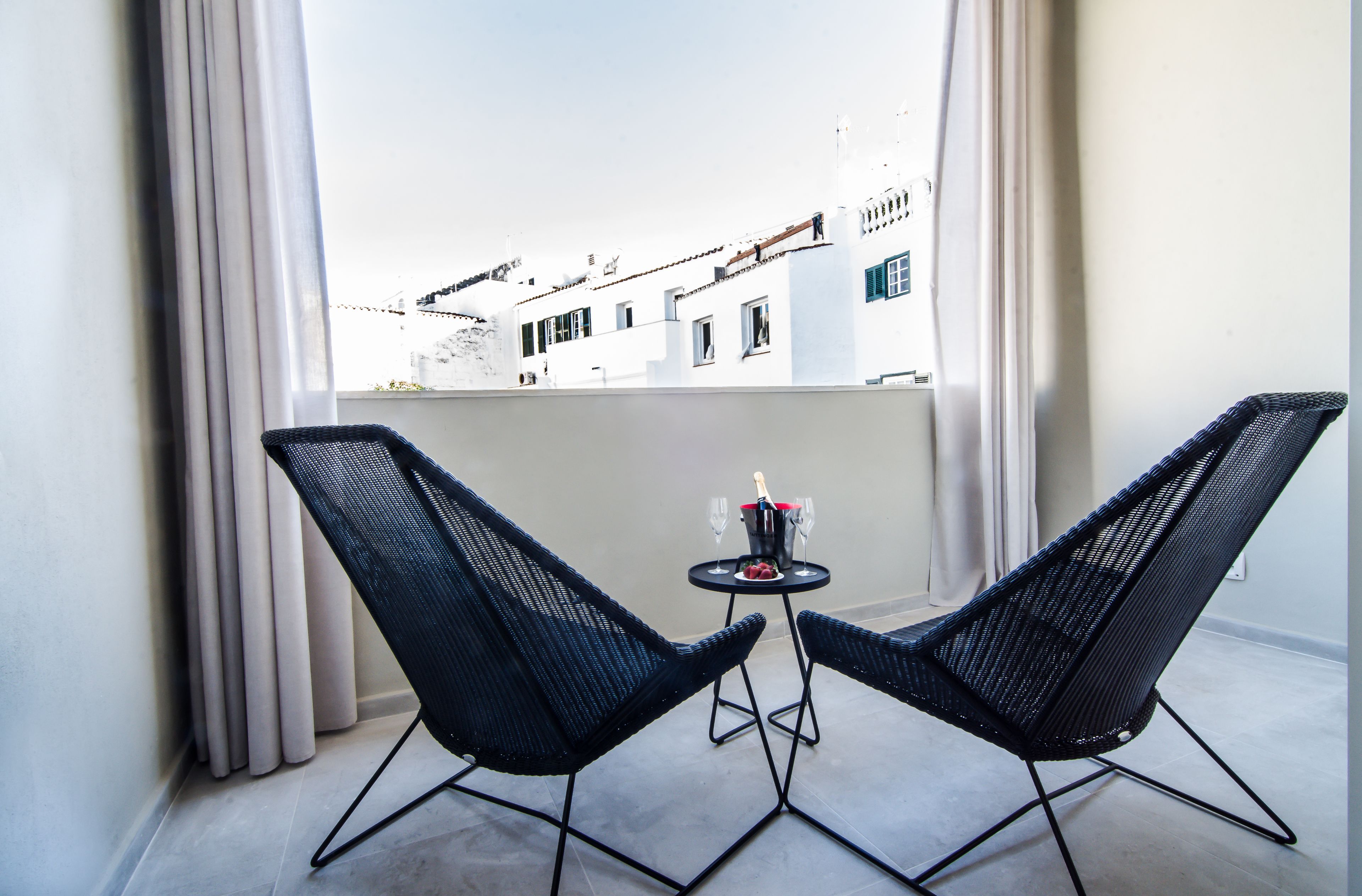 Ein Balkon mit zwei schwarzen Stühlen, einem kleinen Tisch mit einer Flasche und Gläsern sowie Blick auf Gebäude durch transparente Vorhänge.