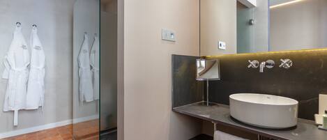 Un baño moderno con un espejo grande, un lavabo redondo y dos batas blancas colgadas en una puerta de cristal.