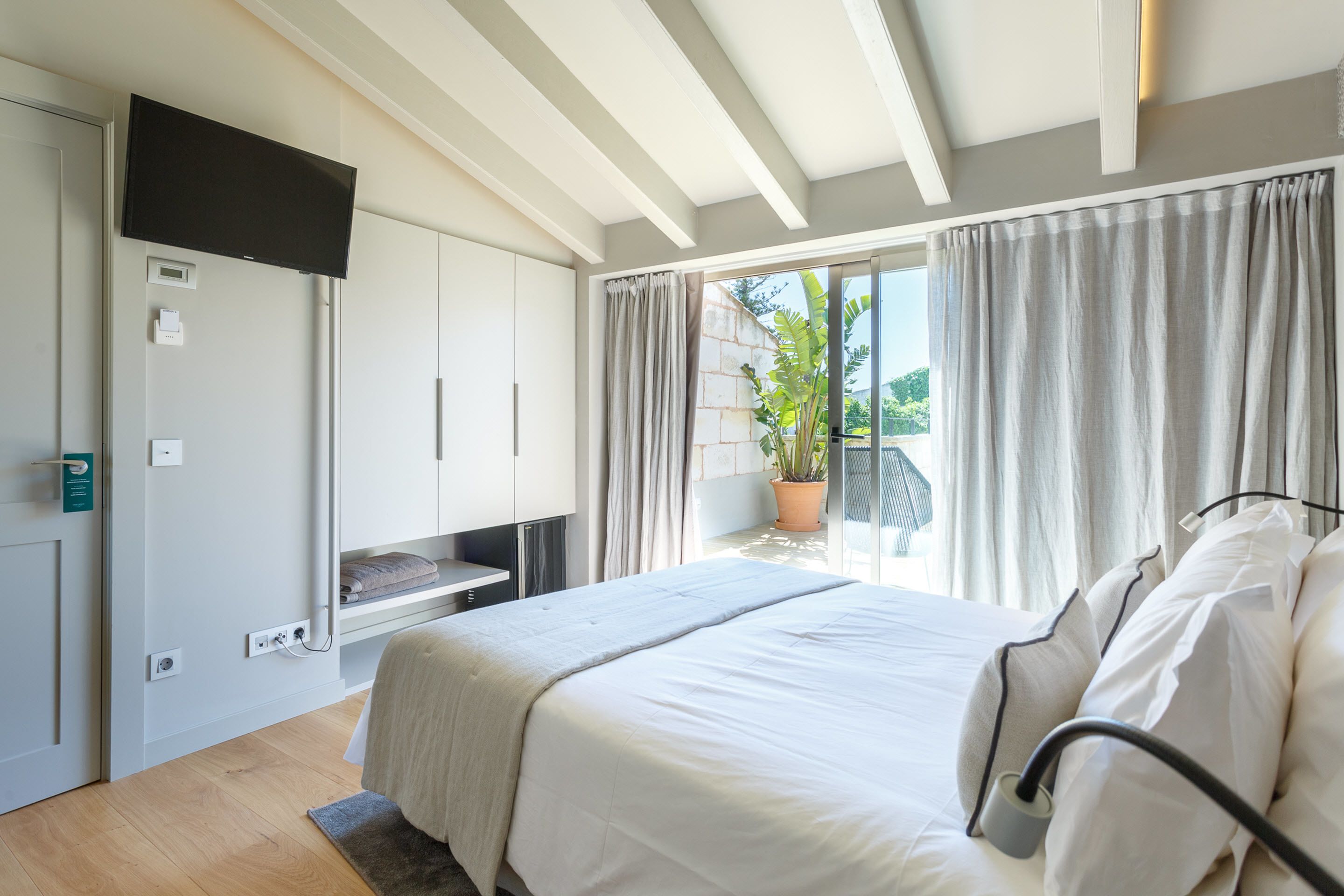 A modern bedroom with a large bed, a TV mounted on the wall, and a balcony with curtains.