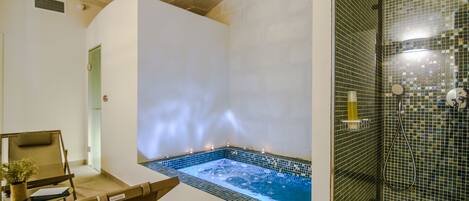 A spa room with a hot tub, a seating area with a chair and a small table, and a shower area with a glass door.