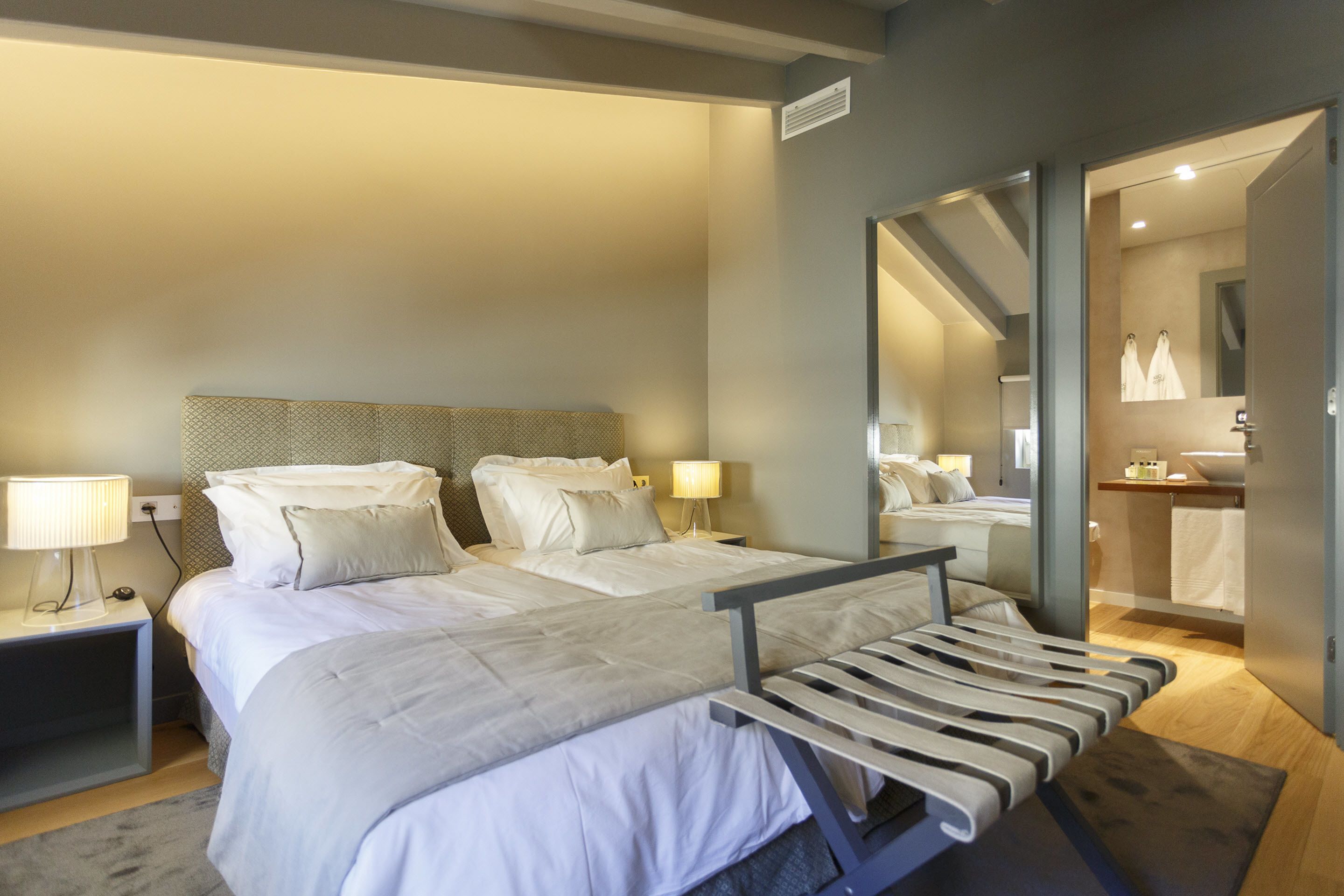 Habitación de hotel moderna con una cama grande, mesitas de noche, un espejo y un baño visible a través de una puerta abierta.