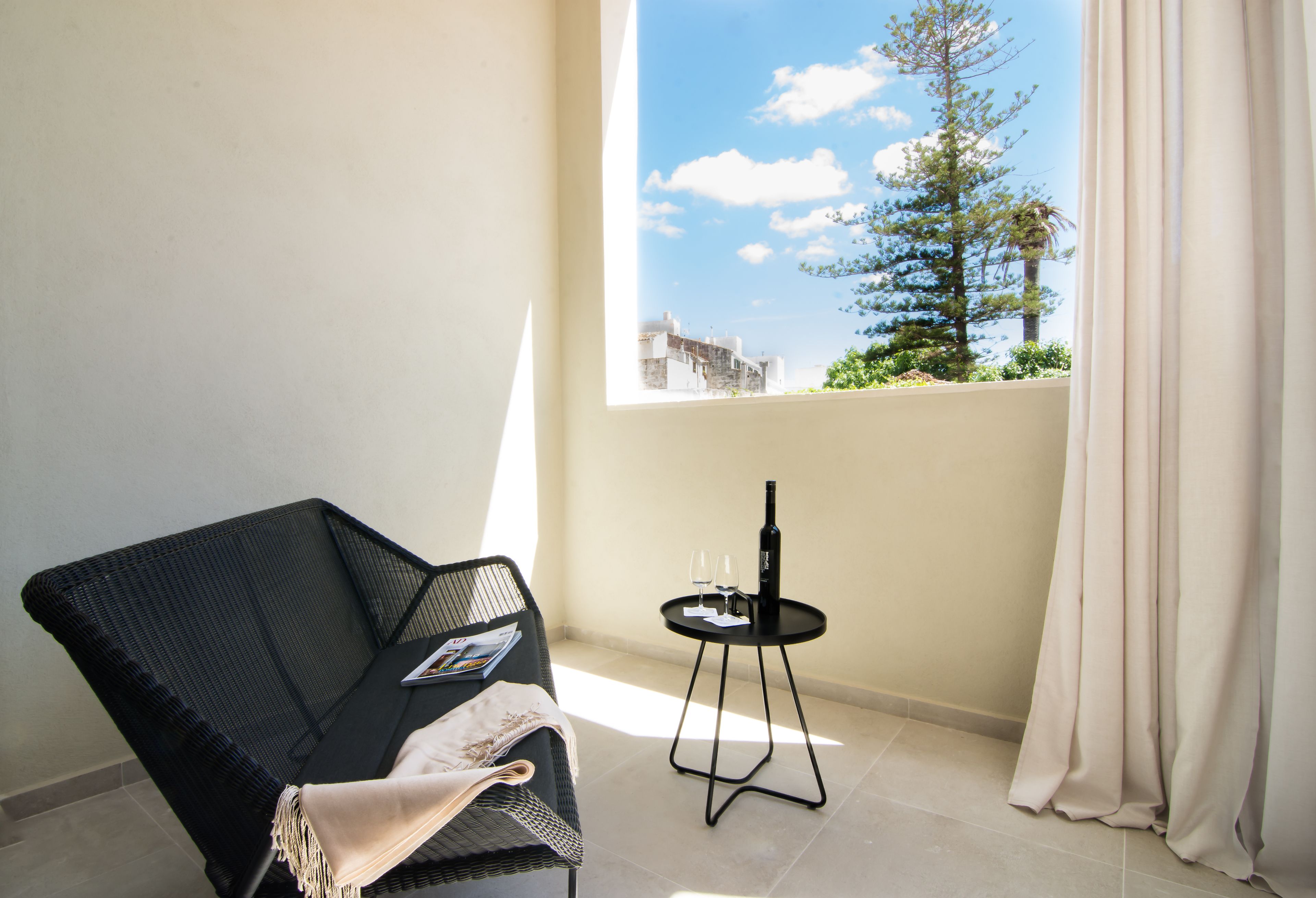Un balcón con una silla de mimbre, una mesita redonda con una botella y vasos, y vistas a un árbol y edificios.