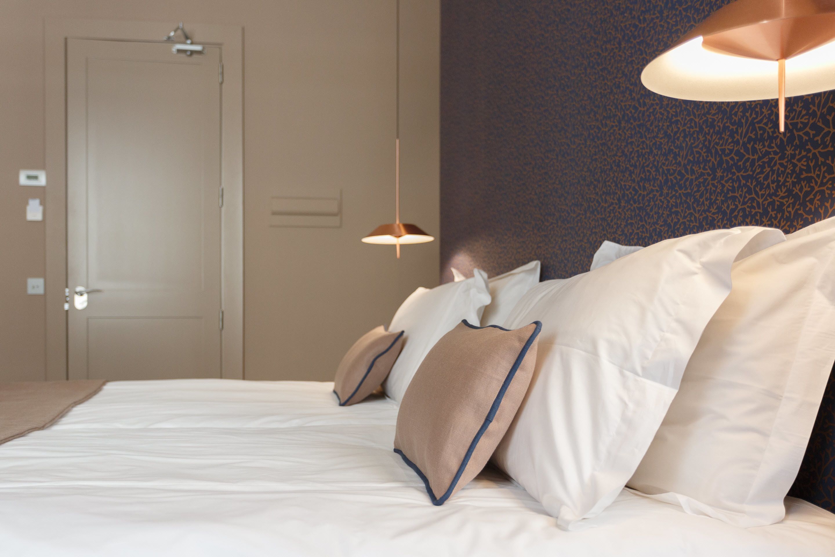 Una cama bien tendida con sábanas blancas y almohadas color beige, una lámpara colgante y una pared texturizada al fondo.