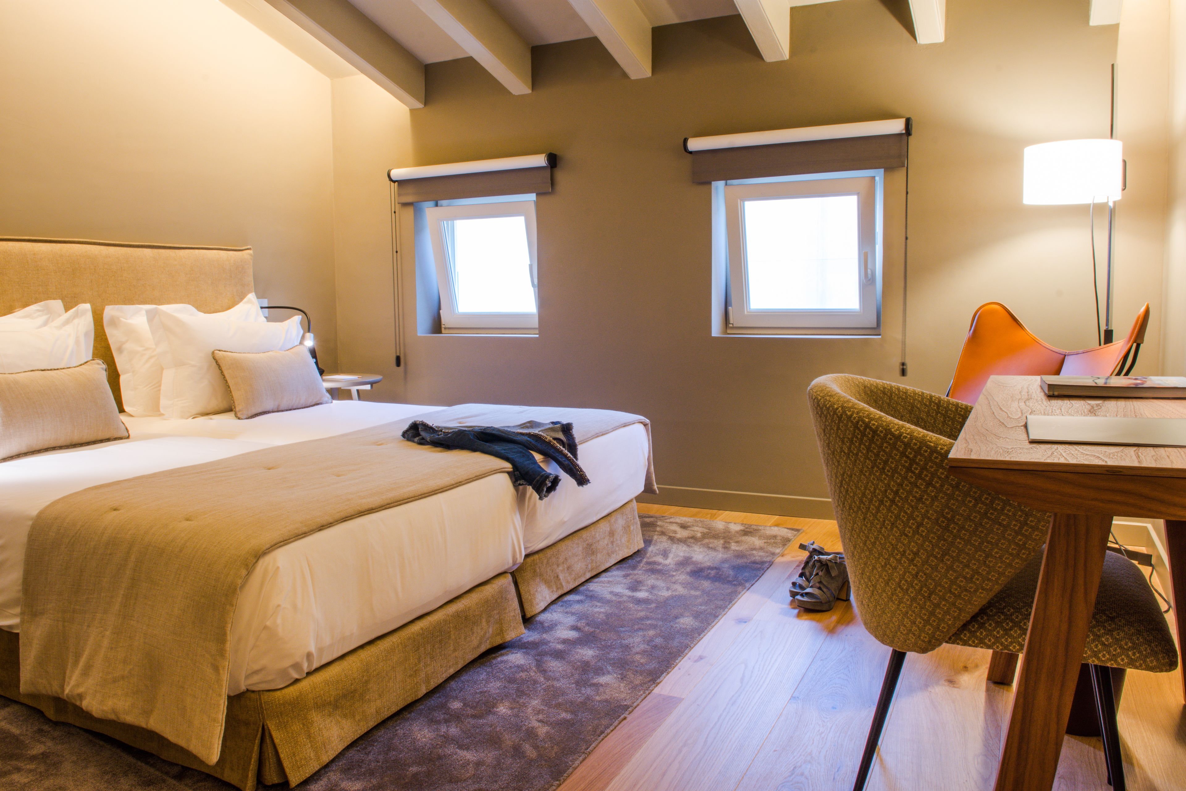 A bedroom with a bed, two windows, a chair, a table, and a lamp.
