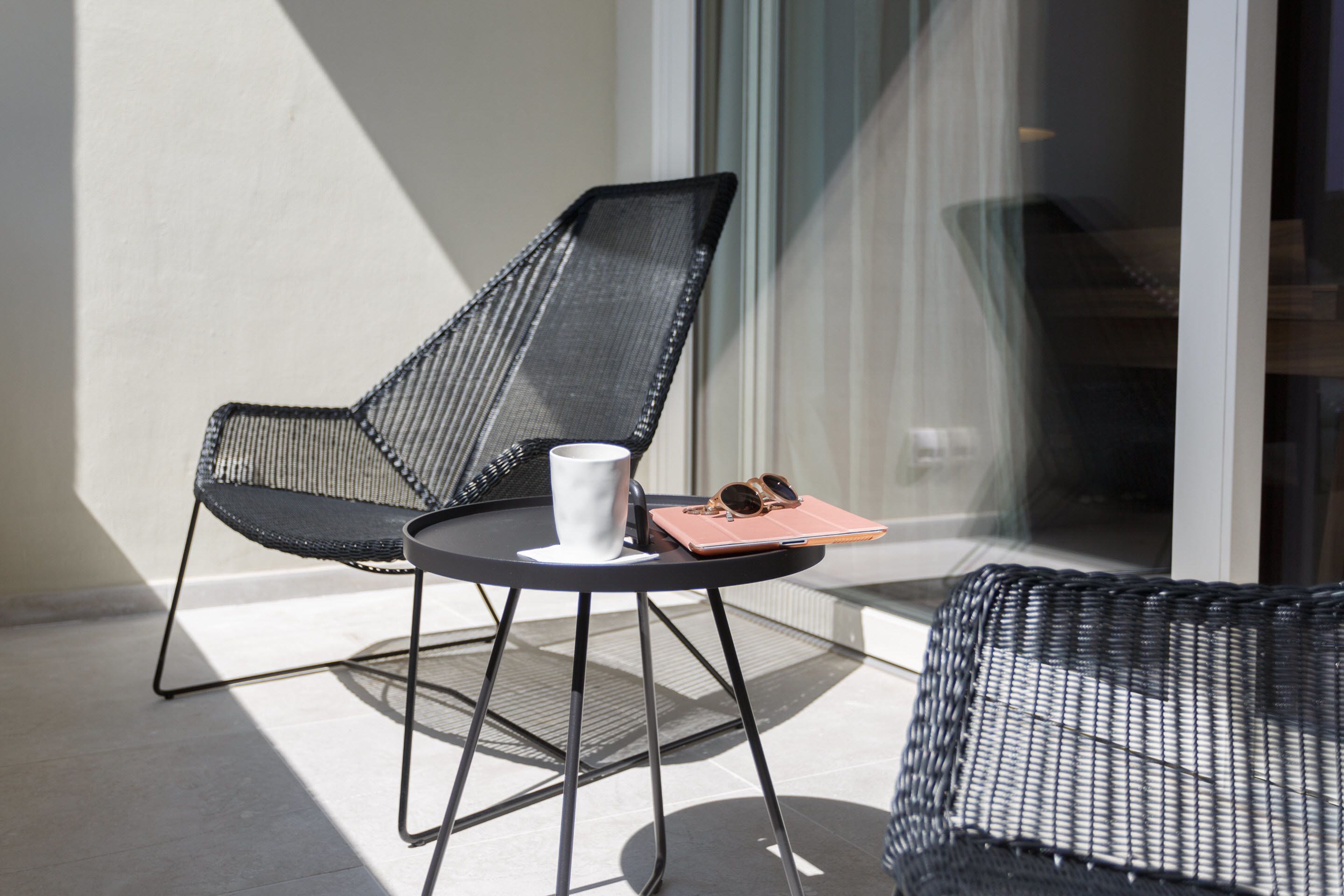 Una silla y una mesa de mimbre negra en un balcón con una pared blanca y una ventana al fondo.