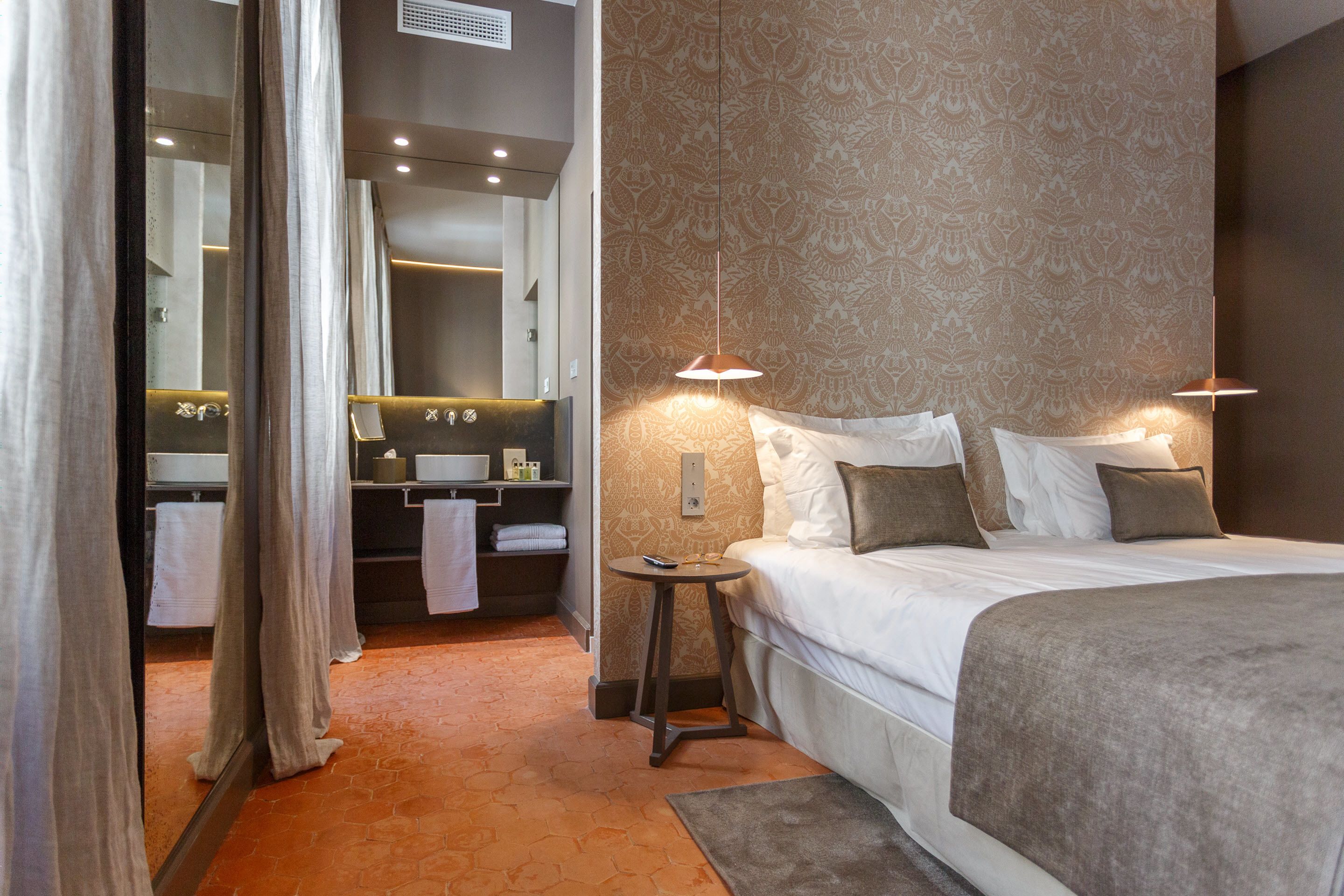 Habitación de hotel con una cama grande, lámparas de noche, una mesita, un espejo y un baño con lavamanos y toallero.