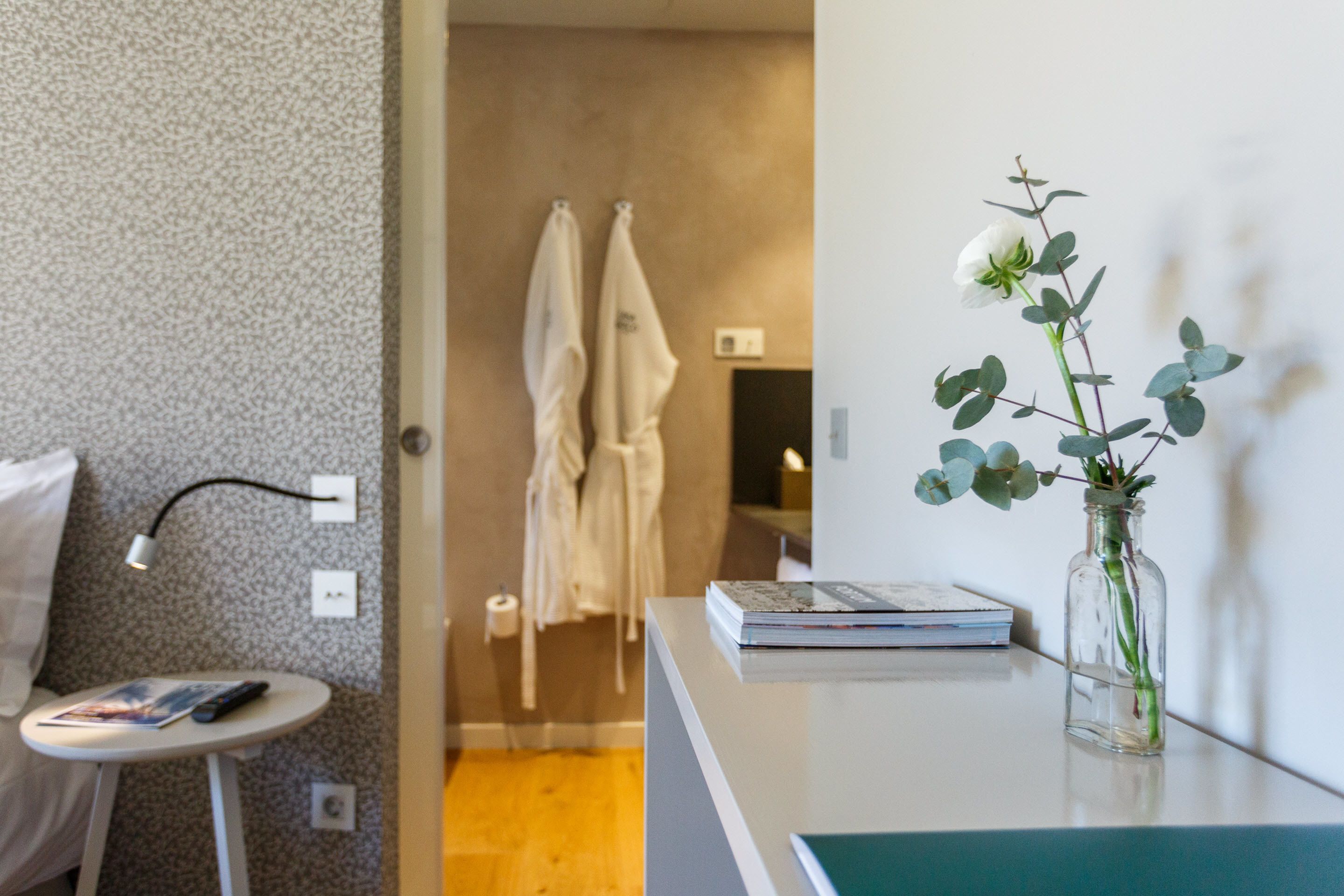 A hotel room with a small table, a vase with a single white flower, and a wall-mounted lamp.