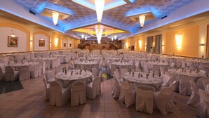 Un salón de banquetes con mesas redondas y sillas, un techo alto con luminarias y un escenario al fondo.