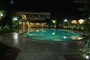 Un área de piscina iluminada con farolas, con un edificio de piedra al fondo y rodeada de árboles.