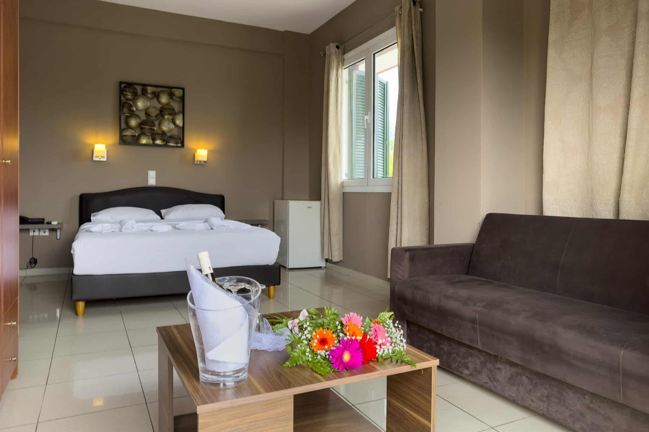 A hotel room with a bed, a bedside table with a glass and flowers, a dark sofa, a wall-mounted light fixture, and a window with curtains.