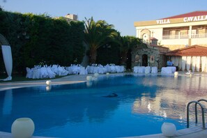 Una piscina con sillas y mesas blancas, rodeada de follaje y un edificio que dice "VILLA CAVALLA".