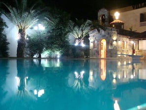 Ein Nachtblick auf einen Swimmingpool mit Palmen und einem Gebäude mit einer beleuchteten Kuppel und einem Rundbogenportal.