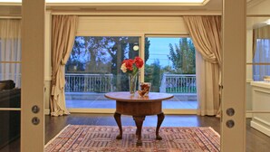 A room with a large sliding glass door leading to a balcony, a round wooden table with a vase of flowers, and a patterned rug on the floor. - Villa Metaxas (Malevizi)