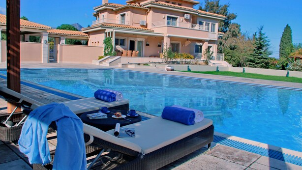 A poolside area with lounge chairs, a table with a towel, and a large house in the background. - Villa Metaxas (Malevizi)