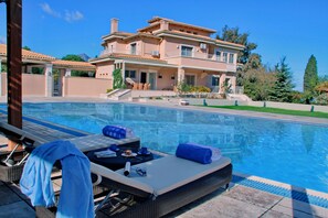 A poolside area with lounge chairs, a pool, and a large house in the background. - Villa Metaxas (Malevizi)