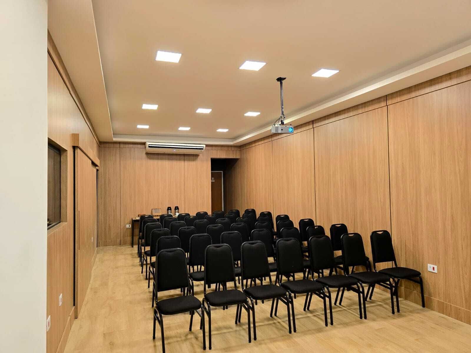 A conference room with rows of black chairs, wooden panel walls, a ceiling projector, and an air conditioning unit.
