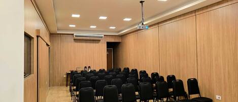 A conference room with rows of black chairs, wooden panel walls, a ceiling projector, and an air conditioning unit.