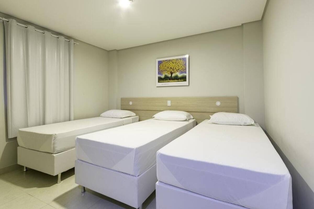Habitación de hotel con tres camas individuales, cabecera de madera, cortina blanca y un cuadro en la pared.