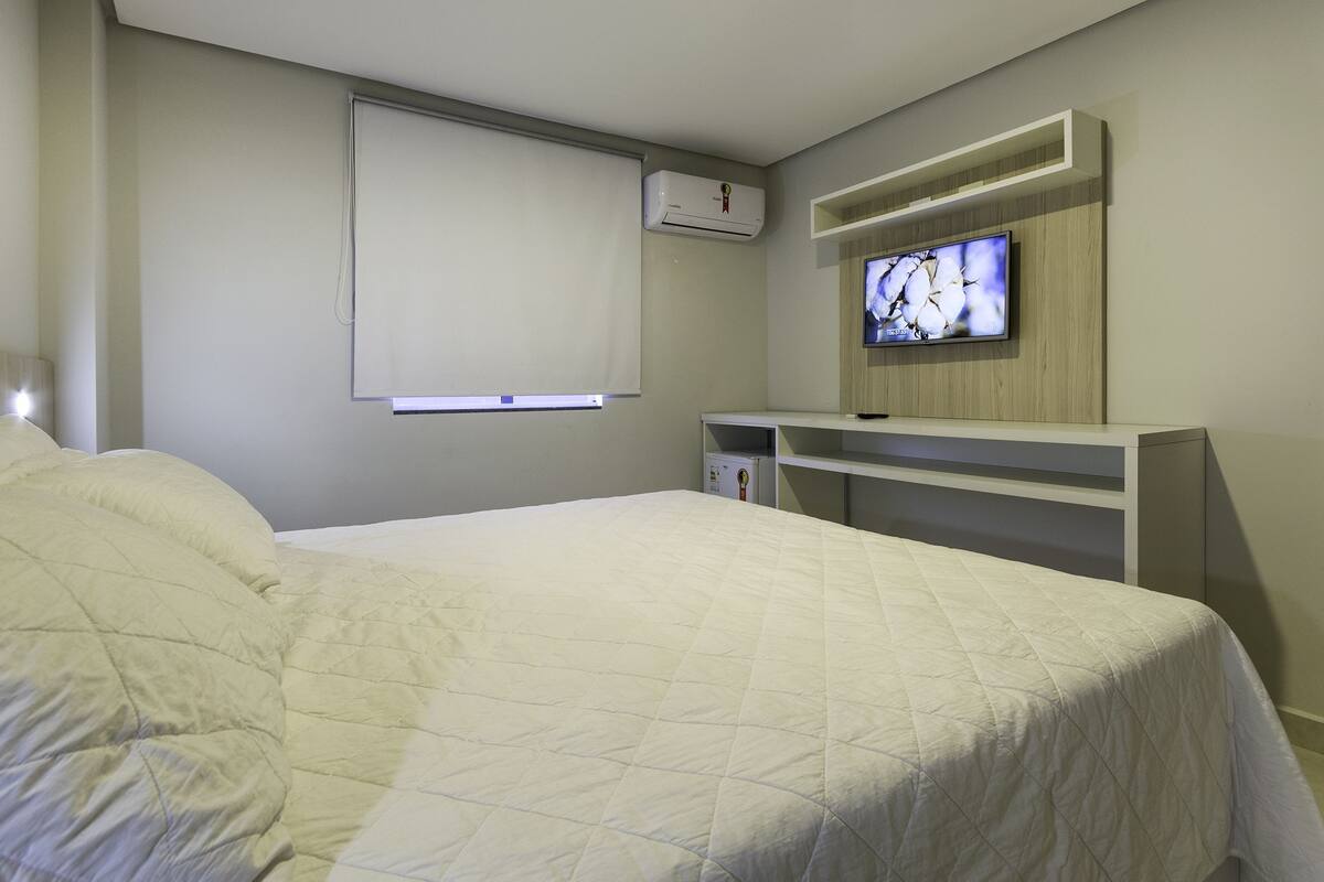 Un dormitorio con cama, televisión montada en la pared, ventana con persianas y estantería empotrada.
