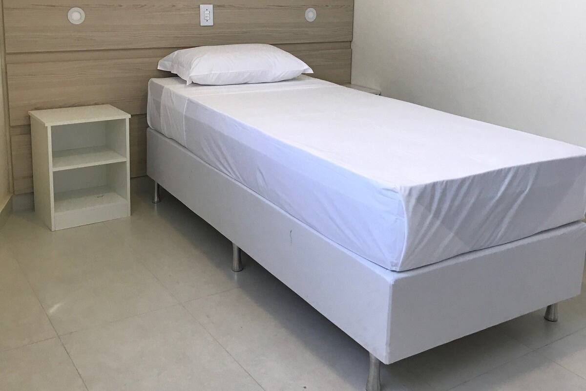 Habitación individual con una cama de una sola plaza, cabecera de madera, colchón blanco, almohada blanca y una mesita de noche pequeña y blanca.