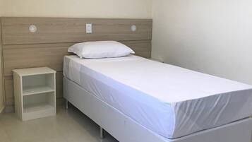 Habitación individual con una cama de una sola plaza, cabecera de madera, colchón blanco, almohada blanca y una mesita de noche pequeña y blanca.