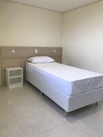 Quarto individual com uma cama de solteiro, cabeceira de madeira, colchão branco, travesseiro branco e mesa de cabeceira pequena e branca.