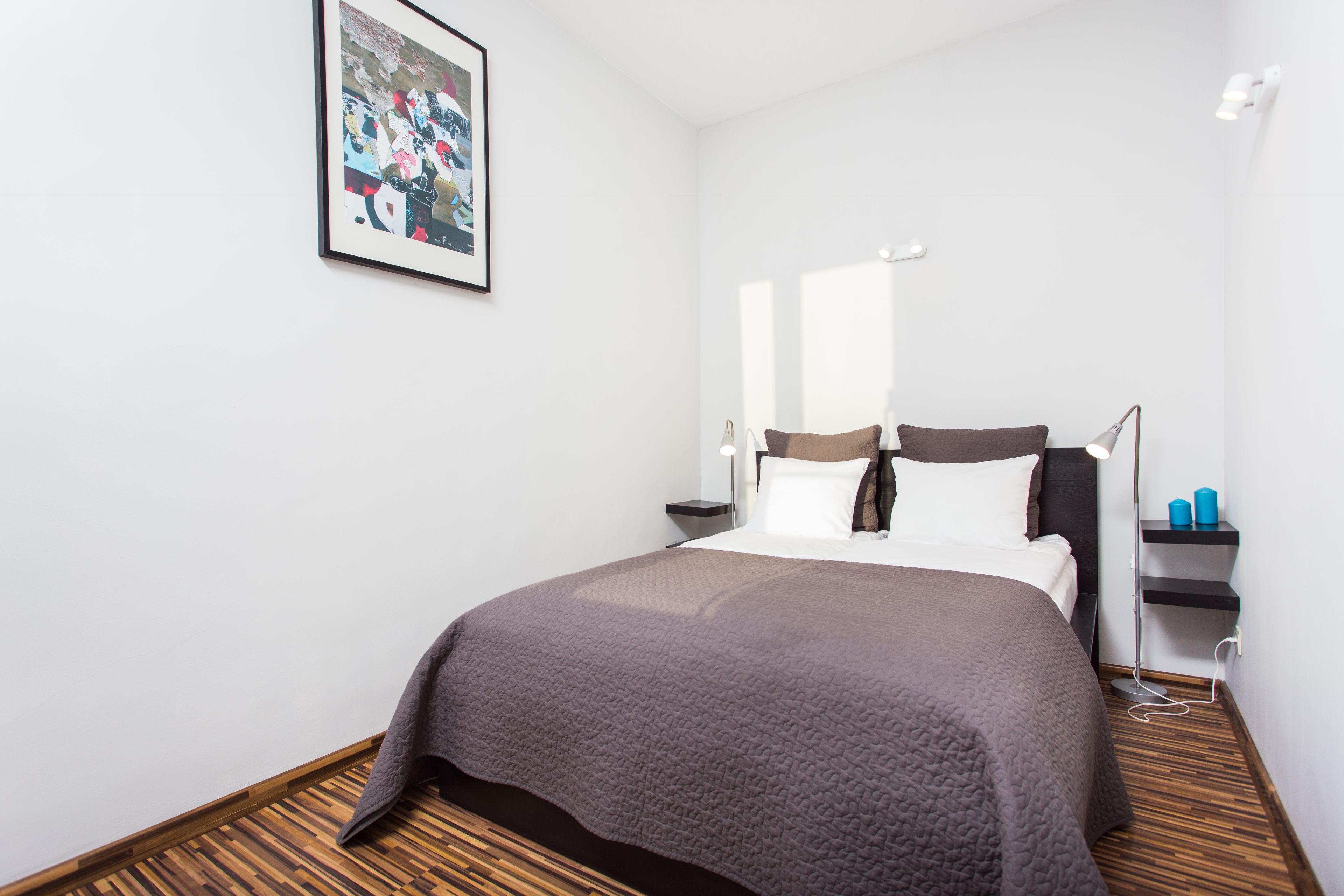 Una cama bien hecha con una manta marrón, una lámpara de noche y un cuadro en la pared.
