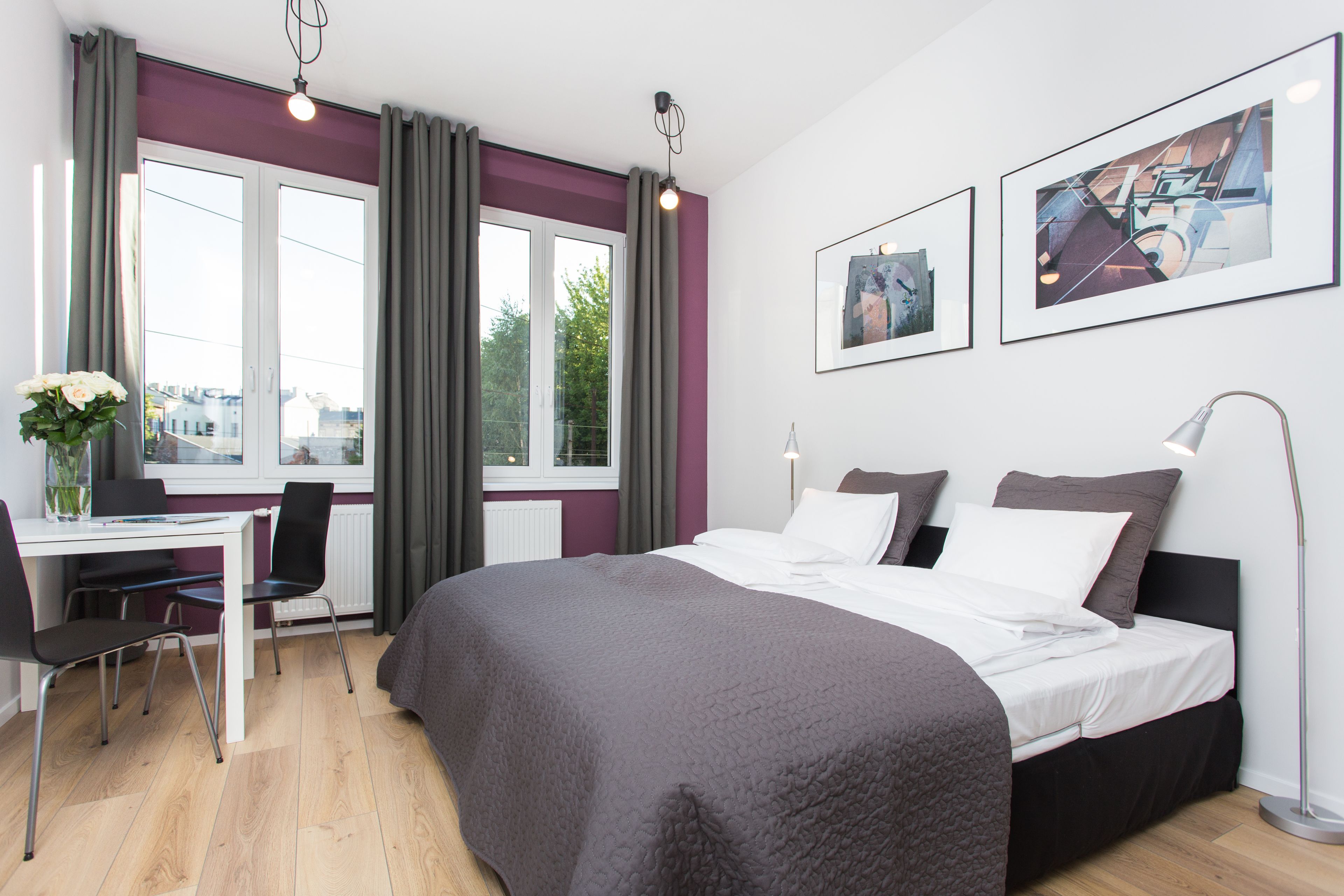 A modern hotel room with a large bed, a desk with a chair, and two windows with curtains. There are two framed pictures on the wall.