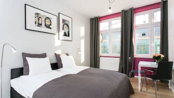 Una habitación de hotel moderna con una cama grande, un escritorio y una silla. Hay dos cuadros en la pared, una ventana con cortinas y una lámpara en la mesita de noche.