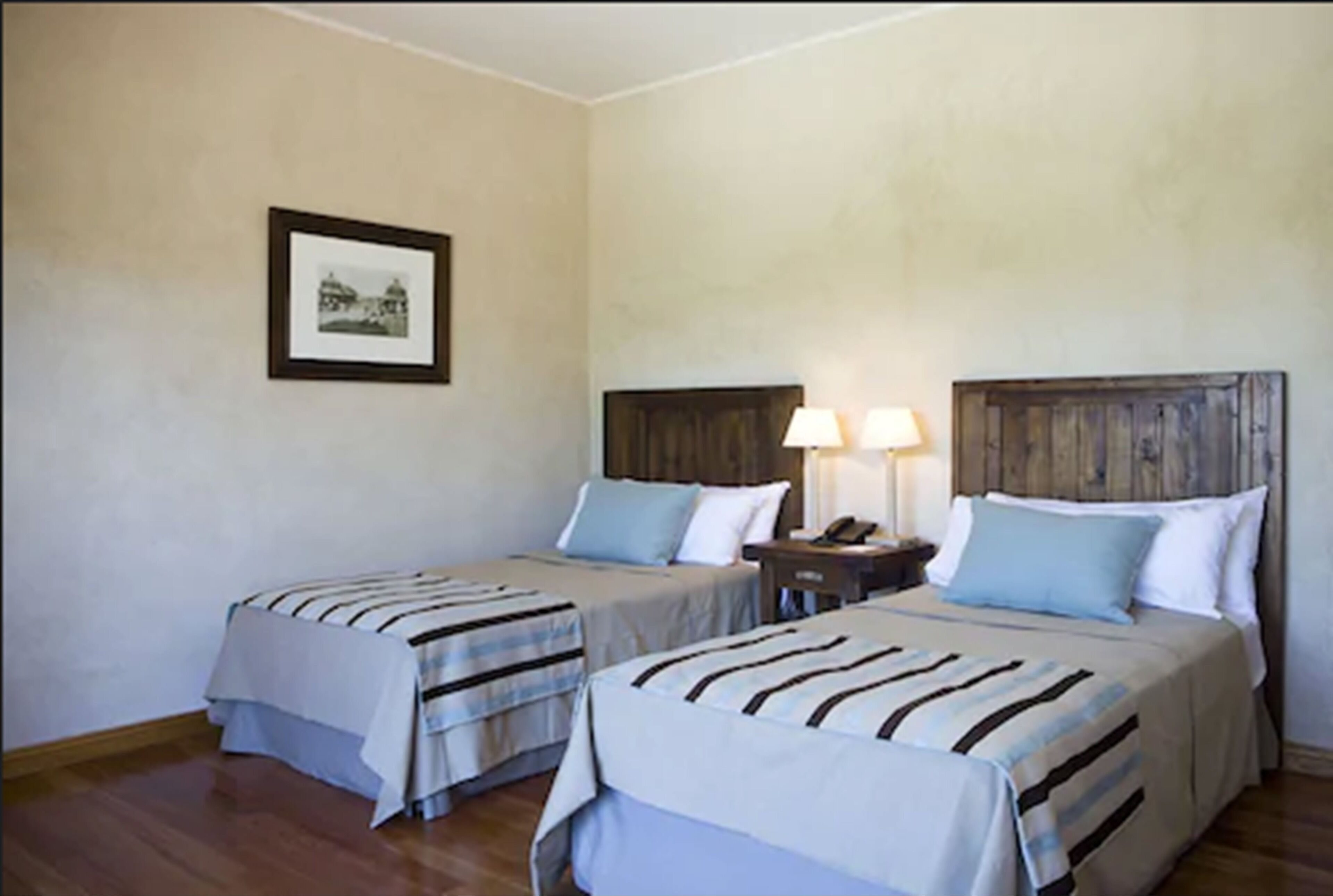 Habitación de hotel con dos camas, un cuadro enmarcado en la pared y una mesita de noche con una lámpara.