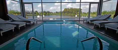A swimming pool with clear blue water, surrounded by lounge chairs and large windows offering a view of a lake and greenery.