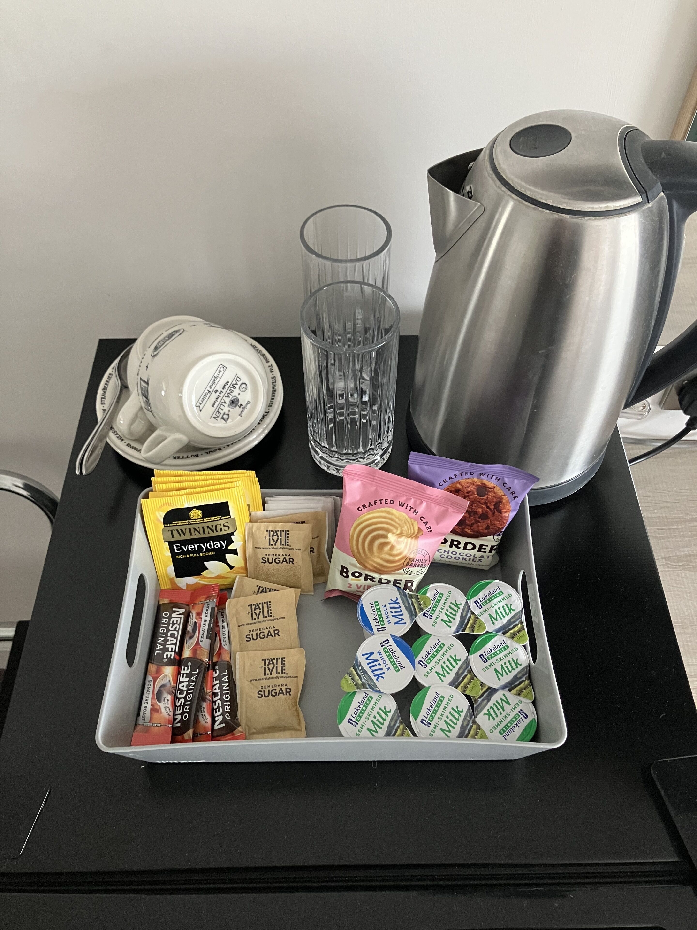 Una bandeja con cafetera, una taza, una tetera y diferentes alimentos, entre ellos café instantáneo, sobres de azúcar y cartones de leche.