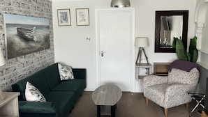 A living room with a green sofa, a small round table, a floral armchair, a lamp, and a painting of a boat on the wall. - Anvershiel House (Portrush)