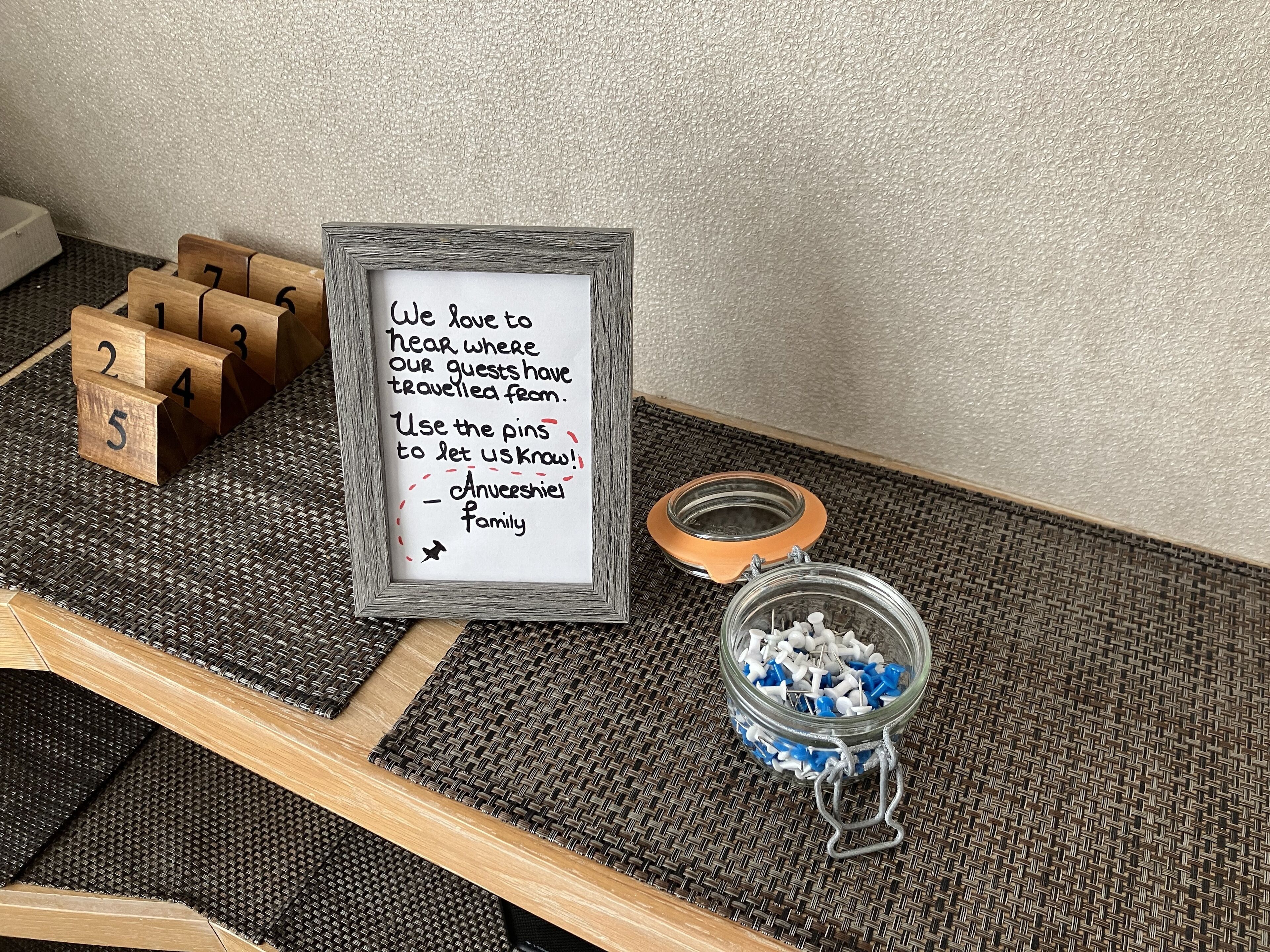 A framed sign with a message about guests' travel history, wooden blocks with numbers, and a jar of pins.