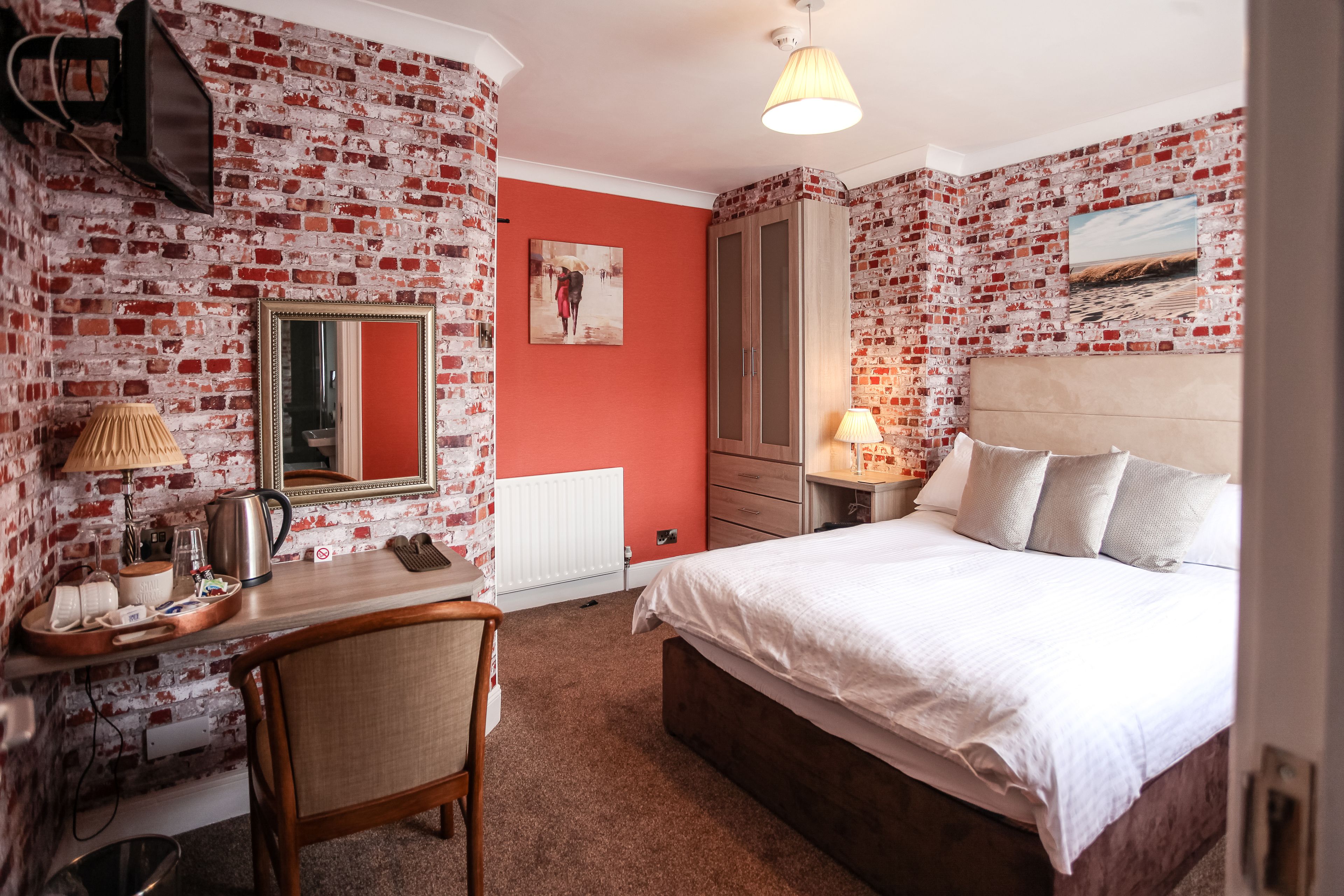 A room with a brick-patterned wall, a bed with white bedding, a wooden desk with a mirror, a chair, and a radiator.