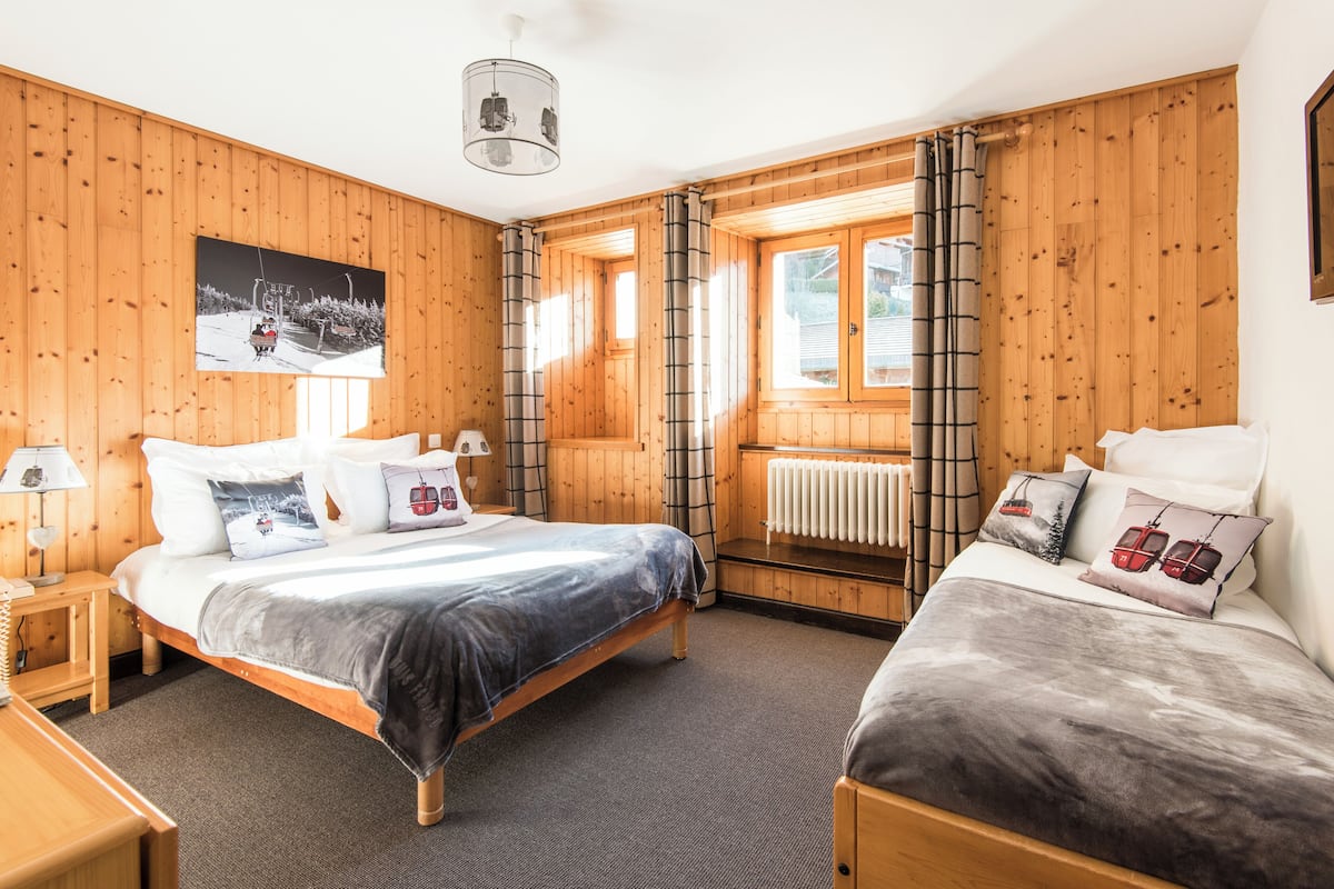 A wooden-paneled bedroom with a bed, a smaller bed, a radiator, and a window with curtains.