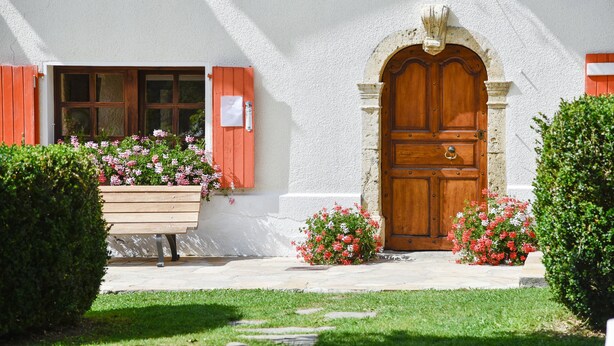 Un edificio con puerta de madera, ventana con contraventanas y un banco lleno de flores.