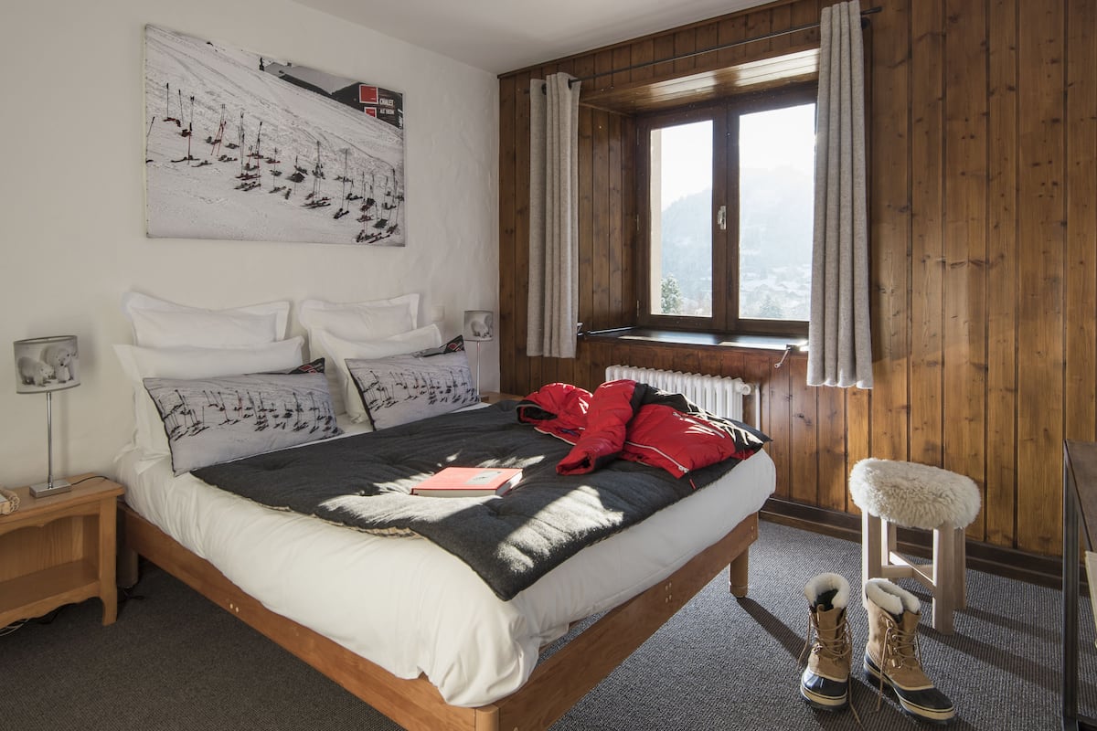 A bedroom with a wooden bed, a window with curtains, a radiator, and a pair of boots on the floor.