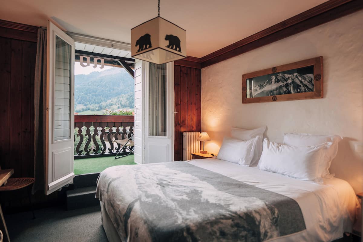 A bedroom with a bed, a nightstand, a lamp, a window with a view of mountains, and a door leading to a balcony.