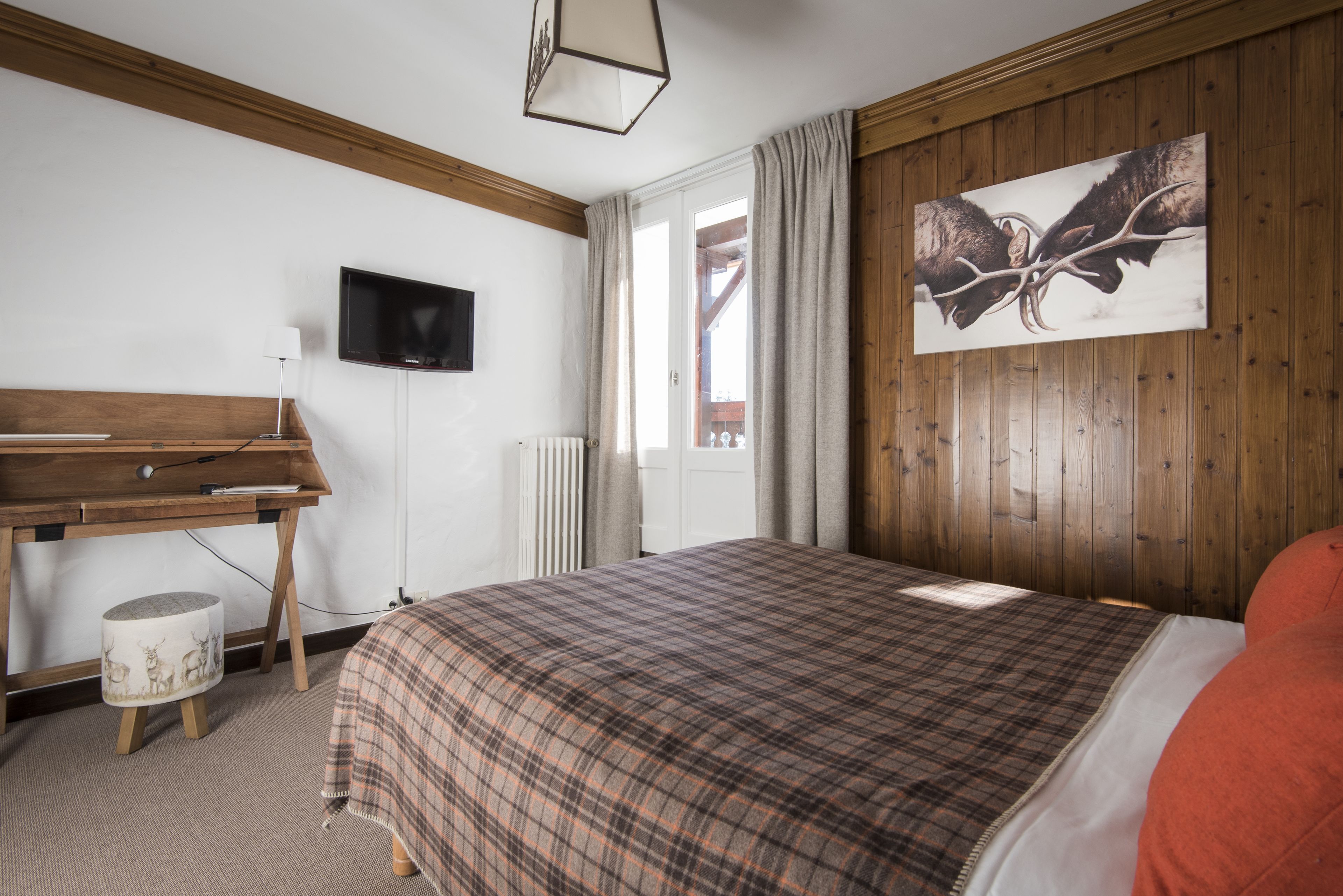 A bedroom with a wooden headboard, a bed with a plaid bedspread, a desk with a lamp, a television on the wall, and a window with curtains.