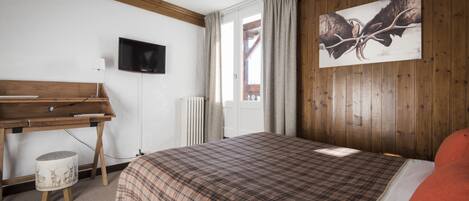 A bedroom with a wooden headboard, a bed with a plaid bedspread, a desk with a lamp, a television on the wall, and a window with curtains.