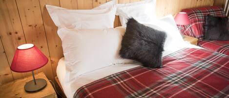 A wooden-paneled room with a bed, a red plaid blanket, white pillows, and a red lamp on a wooden nightstand.