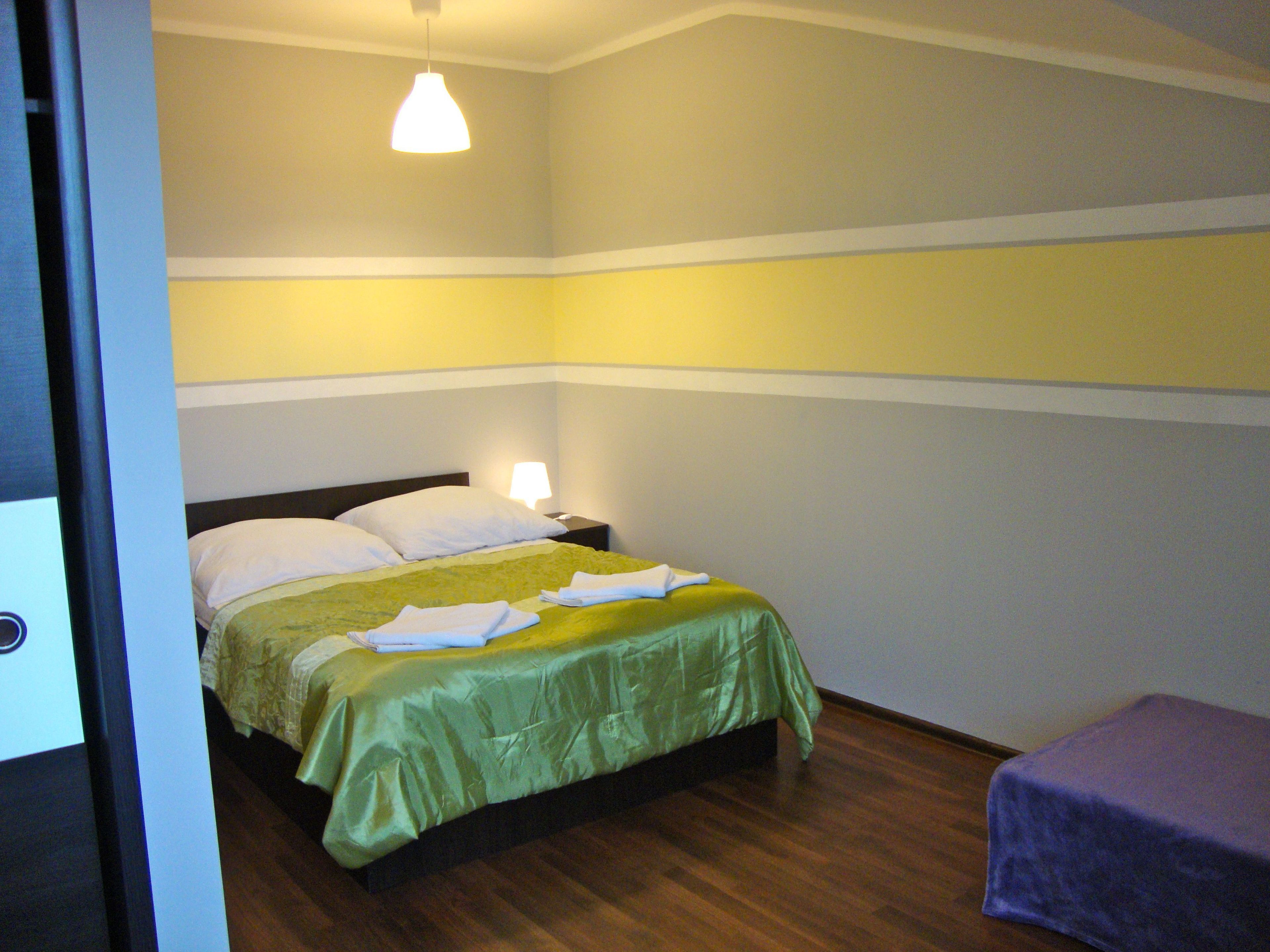 Una cama individual con colcha verde, almohadas blancas y una mesita de noche con lámpara.