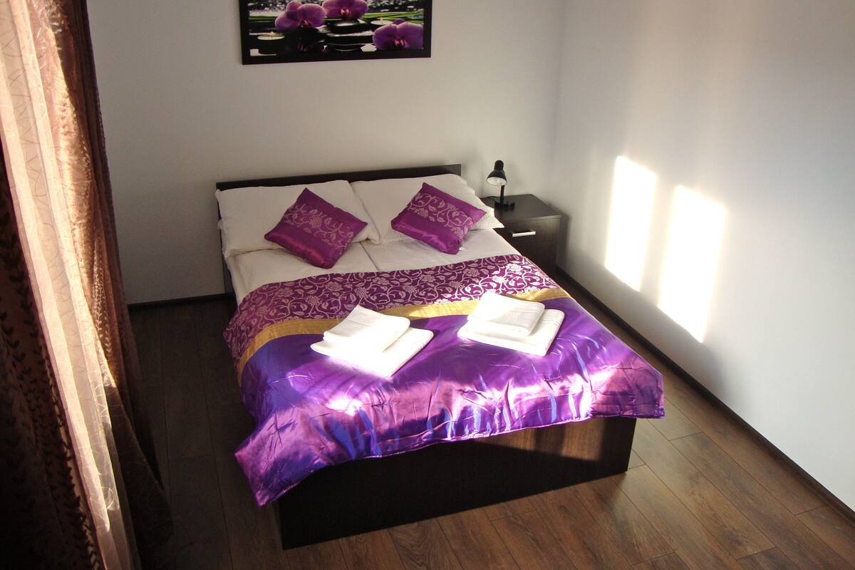 Een netjes opgemaakt bed met paars bedlinnen, een klein nachtkastje met een lamp, en een lijst met een afbeelding erboven.