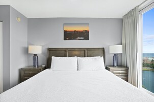 Quarto de hotel com uma cama grande, duas mesas de cabeceira com luminárias, um quadro com uma vista da cidade na parede e vista para um corpo de água pela janela.