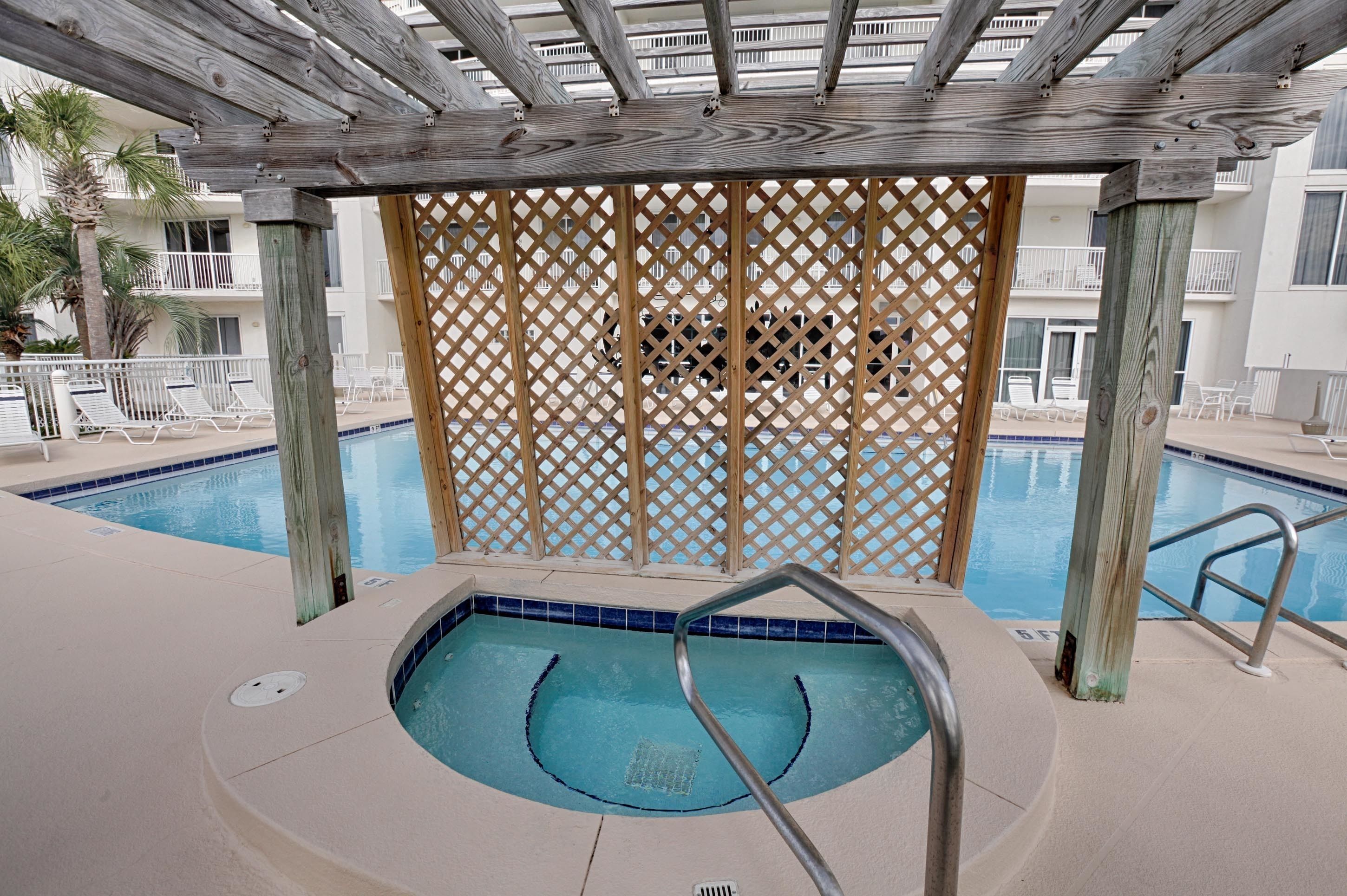Un área de piscina con un pérgola de madera, una piscina con borde de azulejos azules y una valla de madera en el fondo.