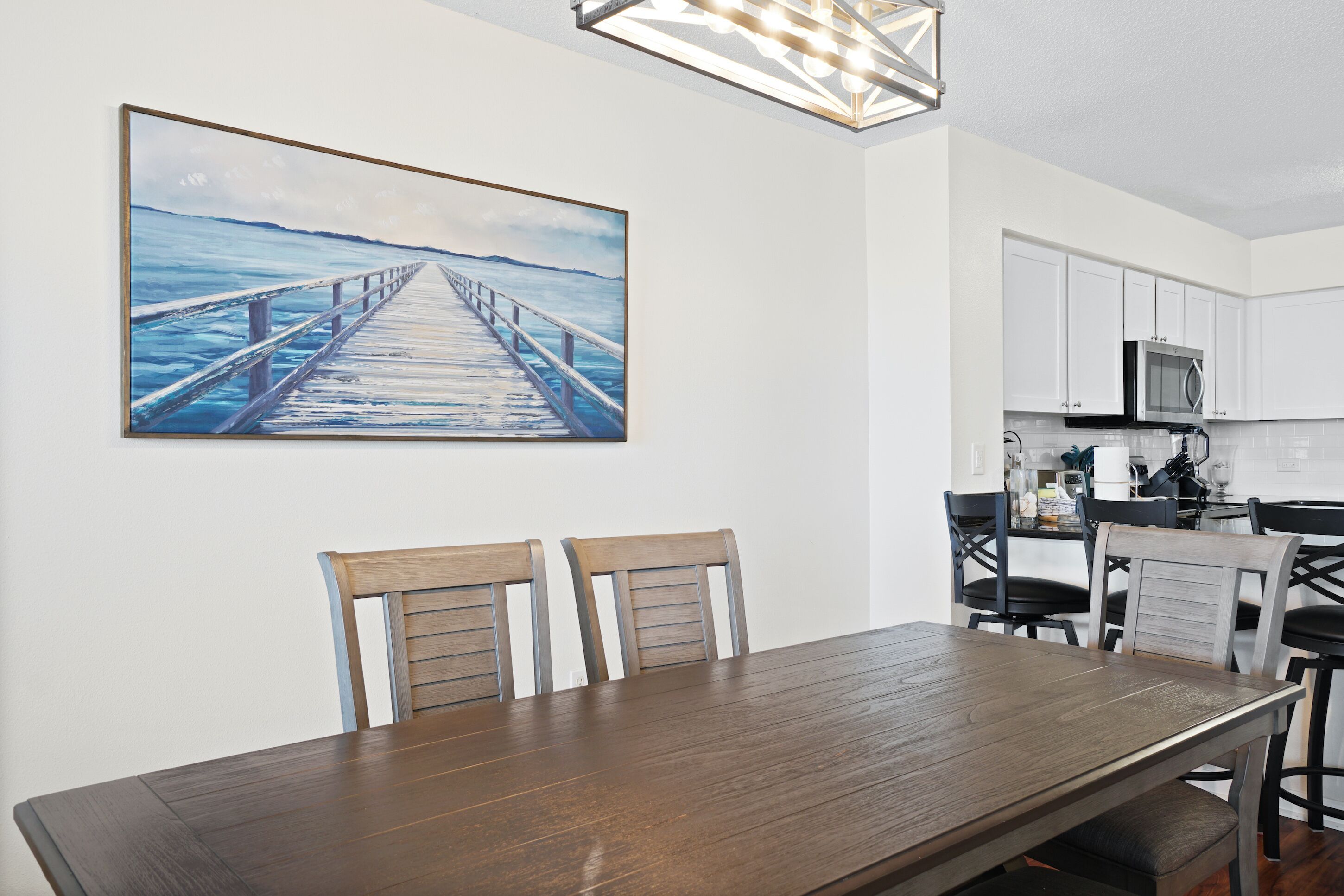 Een eetruimte met een houten tafel en stoelen, een keuken met witte kasten en een magnetron, en een schilderij van een steiger boven het water aan de muur.